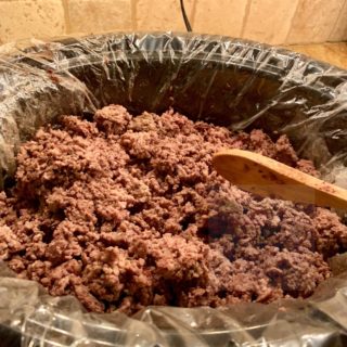 A slow cooker full of browned ground beef being stirred with a wooden spoon