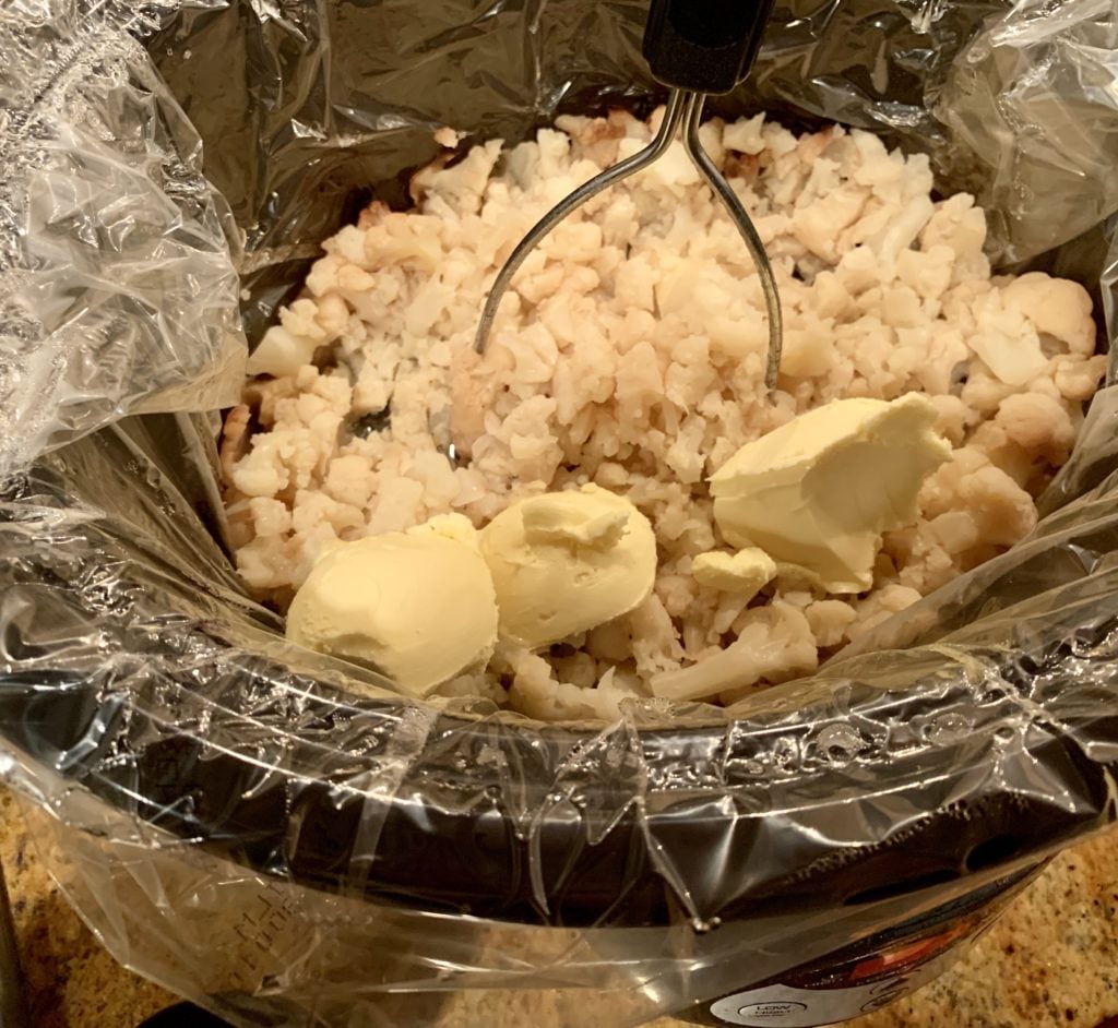 cooked cauliflower in slow cooker with butter substitute and potato masher.