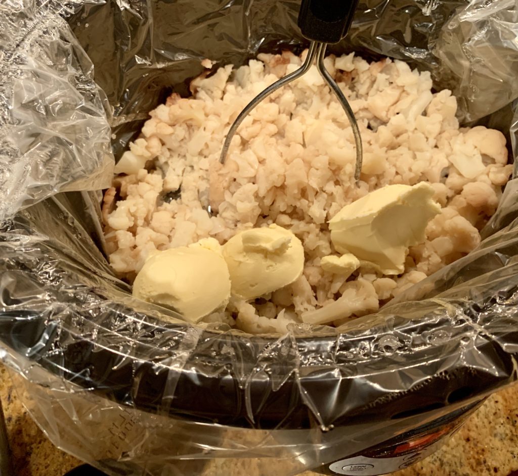 cooked cauliflower in slow cooker with butter substitute and potato masher.