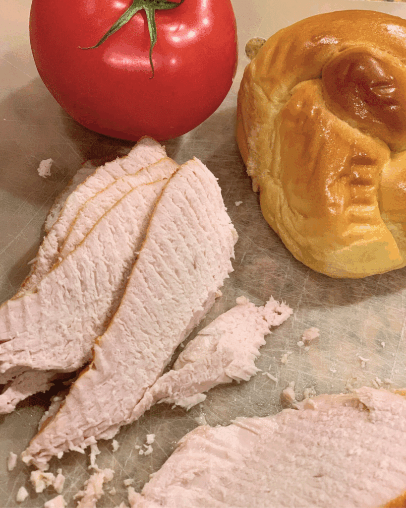 Slices of roasted turkey breast on a cutting board next to a whole tomato and a golden, braided sandwich roll.