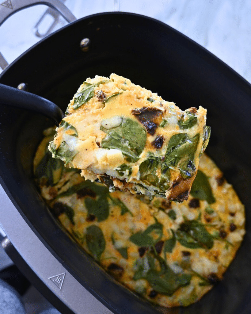 A close-up of a spatula lifting a square piece of spinach and cheese egg casserole from a black slow cooker, with more casserole visible inside the cooker.
