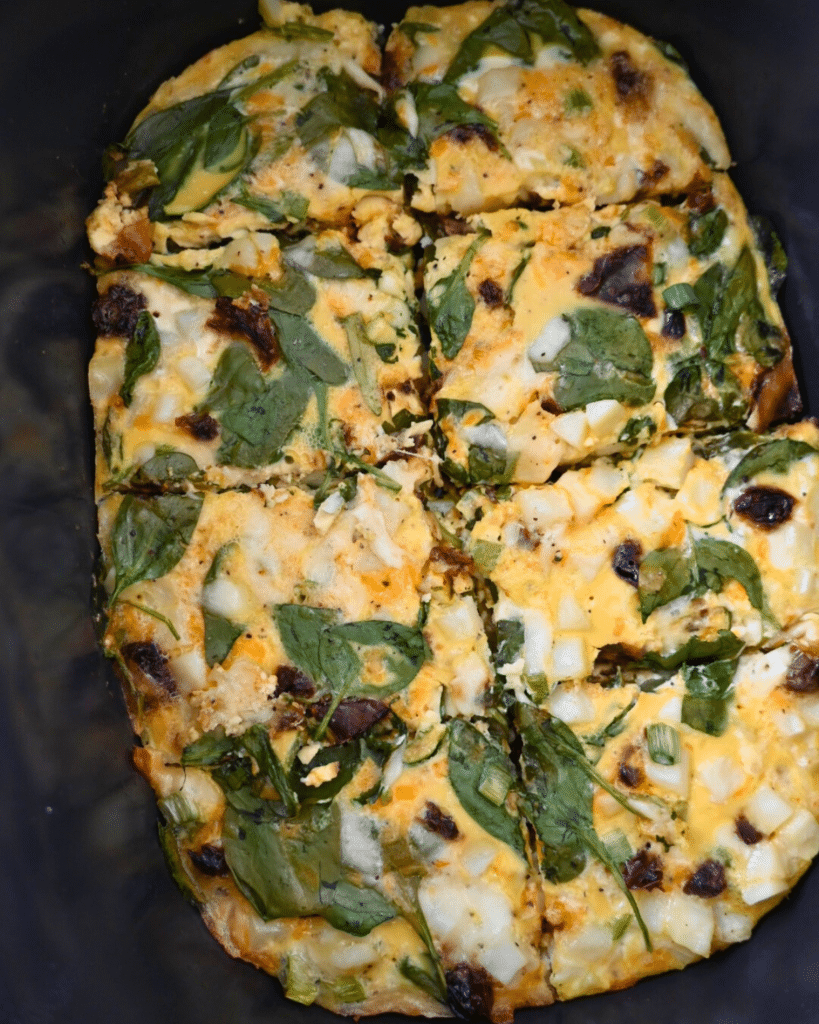 An egg casserole cooked in a slow cooker, topped with spinach leaves, chopped onions, and pieces of sun-dried tomato, with a golden-brown surface.