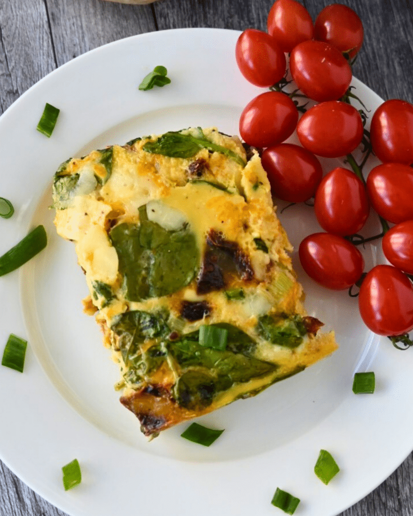 A slice of vegetable frittata with spinach on a white plate, garnished with green onion and served with a bunch of grape tomatoes. The plate is on a rustic wooden surface.
