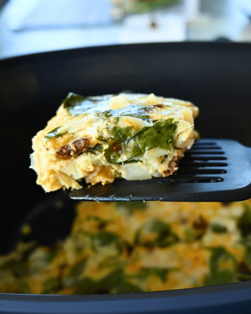 A close-up of a spatula lifting a square piece of spinach and cheese egg casserole from a black slow cooker, with more casserole visible inside the cooker.
