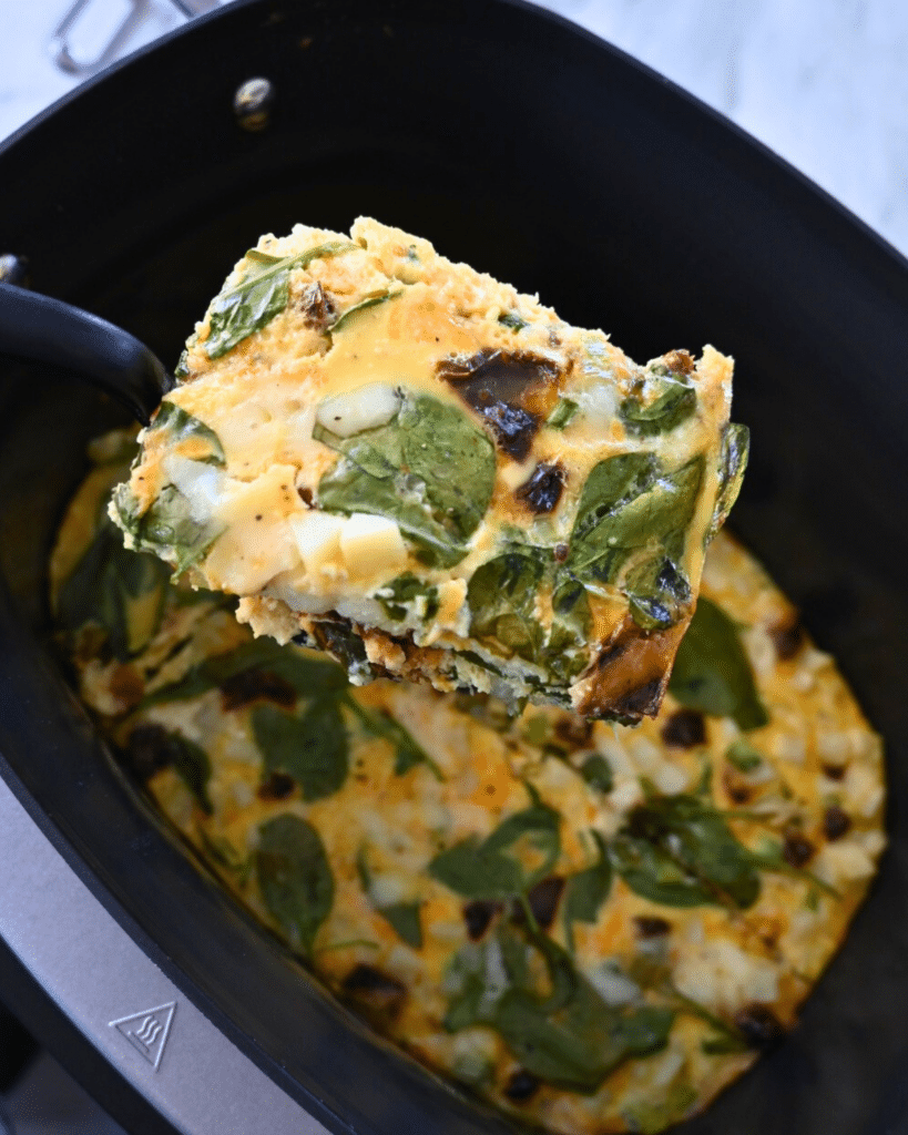 A close-up of a spatula lifting a square piece of spinach and cheese egg casserole from a black slow cooker, with more casserole visible inside the cooker.