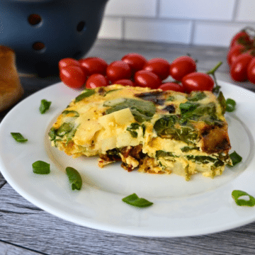 A slice of vegetable frittata with spinach on a white plate, garnished with green onion and served with a bunch of grape tomatoes. The plate is on a rustic wooden surface.