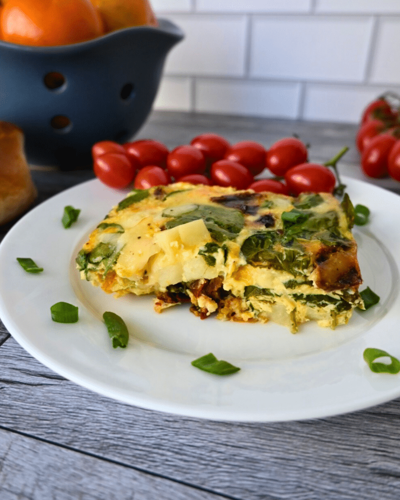 A slice of vegetable frittata with spinach on a white plate, garnished with green onion and served with a bunch of grape tomatoes. The plate is on a rustic wooden surface.