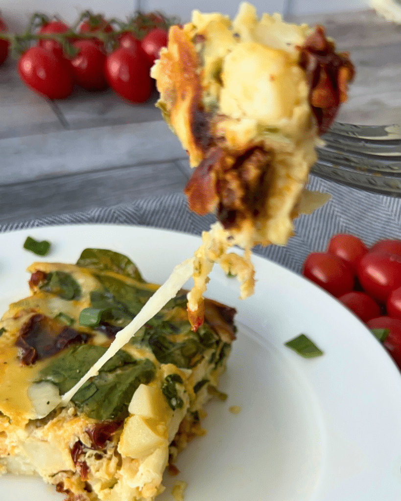 A close-up of a fork holding a cheesy bite of vegetable frittata above a plate, with visible spinach, sun-dried tomatoes, and melted cheese. Fresh cherry tomatoes are in the background.