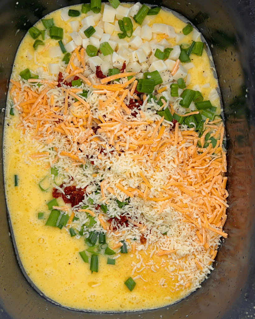 A close-up of a crockpot filled with whisked eggs, shredded cheddar and mozzarella cheese, chopped green onions, sun-dried tomatoes, and diced white cheese, ready to be mixed together.
