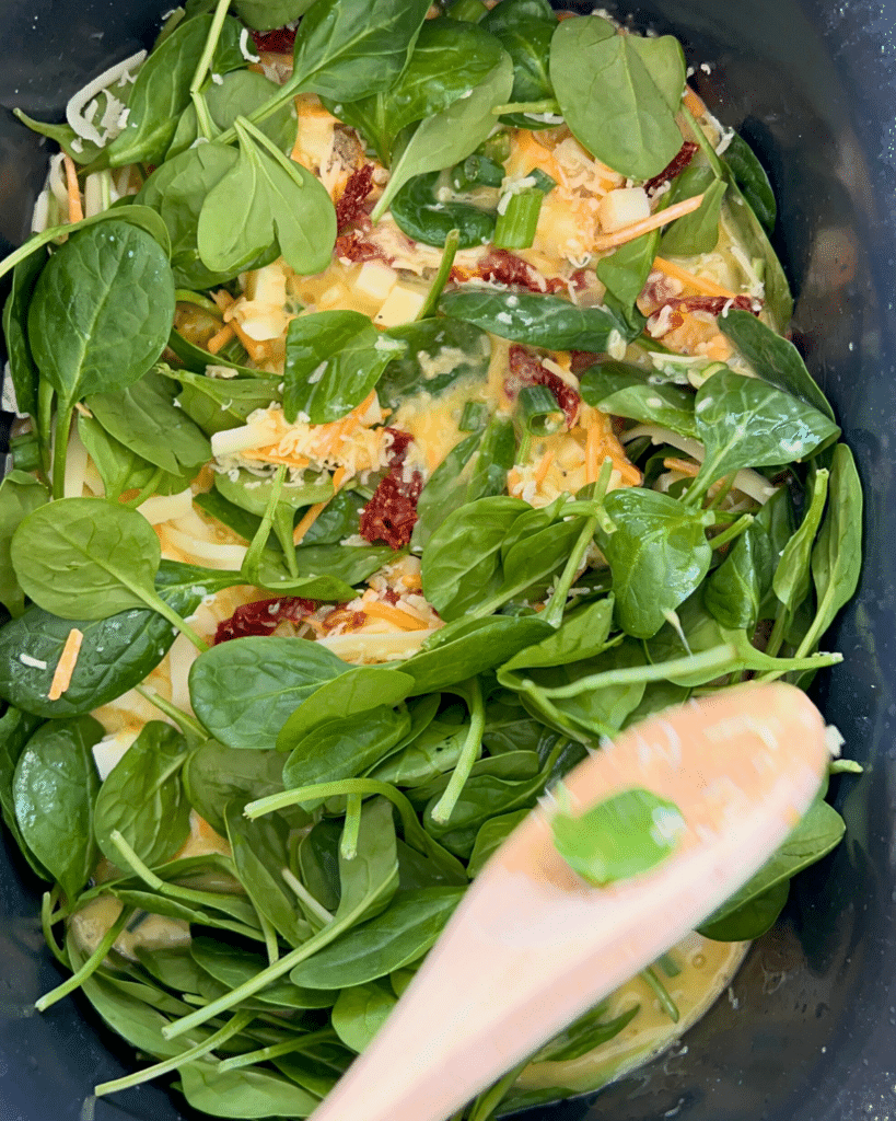 A close-up of a slow cooker filled with whisked eggs, shredded cheddar and mozzarella cheese, chopped green onions, sun-dried tomatoes, and diced white cheese, ready to be mixed together.
