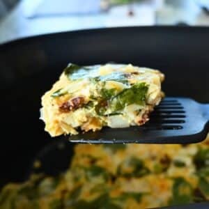 A close-up of a spatula lifting a square piece of spinach and cheese egg casserole from a black slow cooker, with more casserole visible inside the cooker.