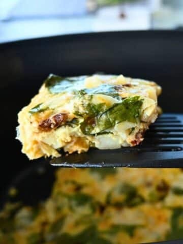 A close-up of a spatula lifting a square piece of spinach and cheese egg casserole from a black slow cooker, with more casserole visible inside the cooker.