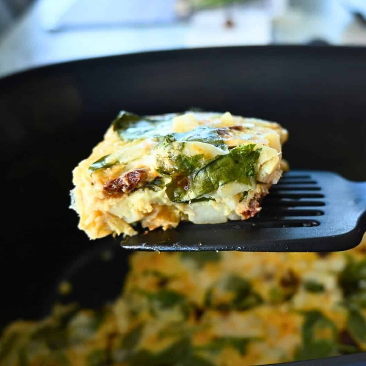 A close-up of a spatula lifting a square piece of spinach and cheese egg casserole from a black slow cooker, with more casserole visible inside the cooker.