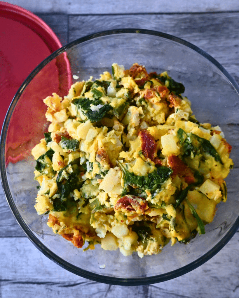 A glass bowl filled with a scrambled leftover Frittata mixture containing potatoes, spinach, and sun-dried tomatoes sits on a wooden surface next to a red lid.