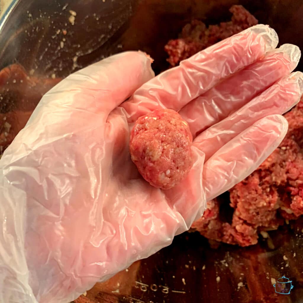 A hand wearing a glove holding a formed meatball with a bowl of the meat mixture in the background