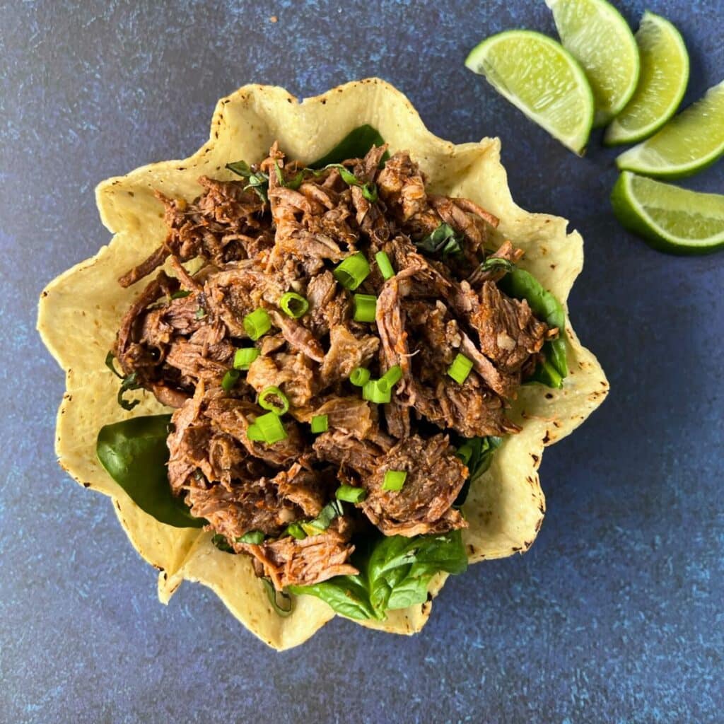 a tortilla bowl filled with shredded beef barbacoa and topped with sliced green onion with lime wedges