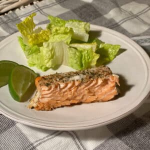 A round white plate with a piece of cooked salmon, lettuce and lime wedges