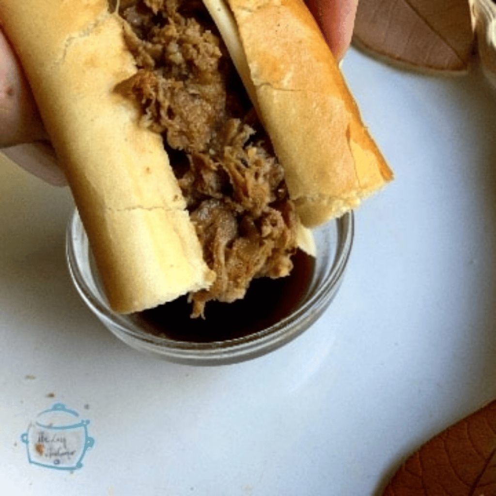a roll filled with shredded beef held over a small glass bowl filled with au just ready to dip