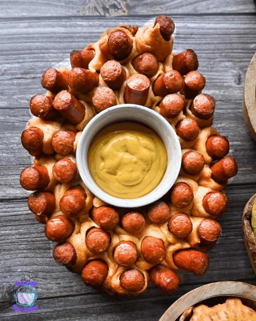An oval-shaped pull-apart bread with mini hot dogs baked in, arranged around a central bowl of yellow mustard dipping sauce, set on a wooden surface.