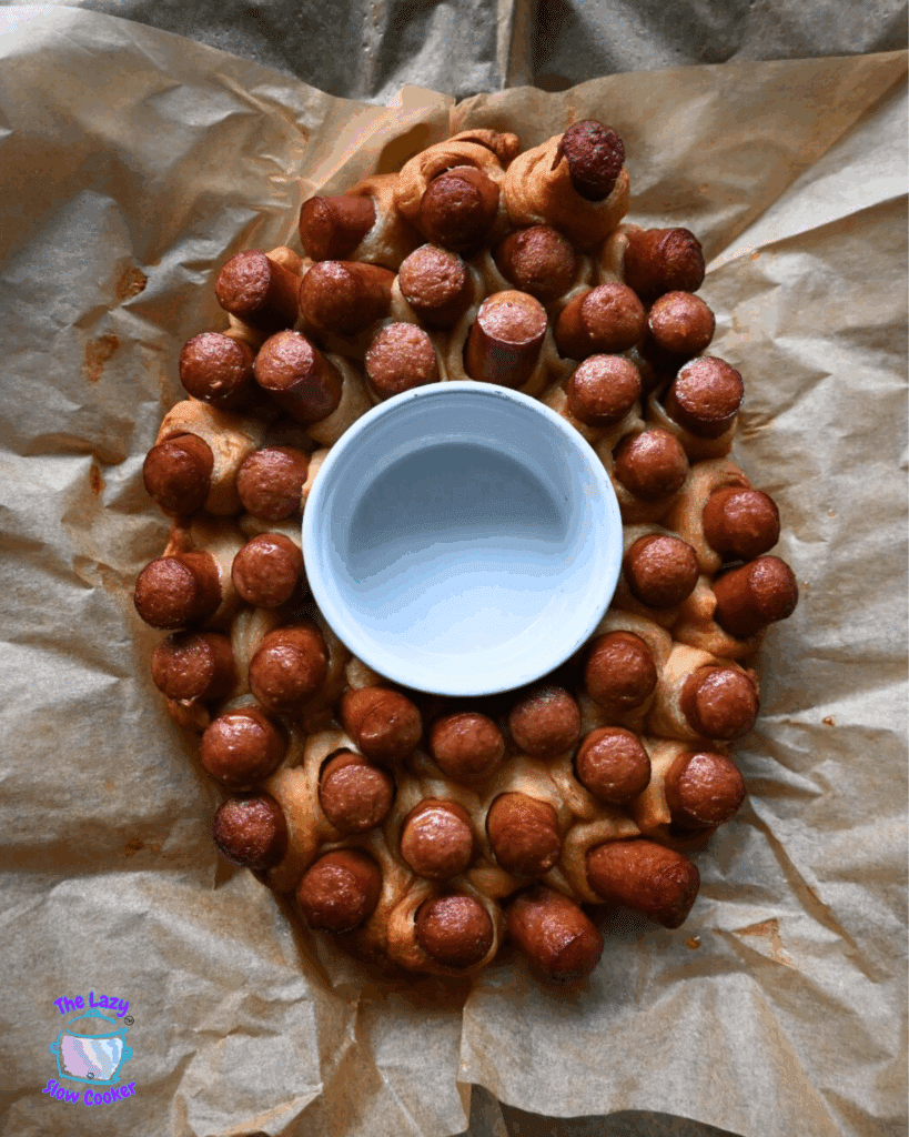 A slow cooked appetizer shaped like a football, made of small hot dogs wrapped in dough, surrounds a white bowl in the center on parchment paper. The Lazy Slow Cooker logo is visible in the lower left corner.