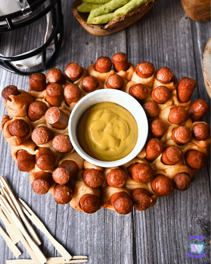Oval slow cooker-shaped pull-apart bread with mini hot dogs baked in, arranged around a central bowl of yellow mustard dipping sauce