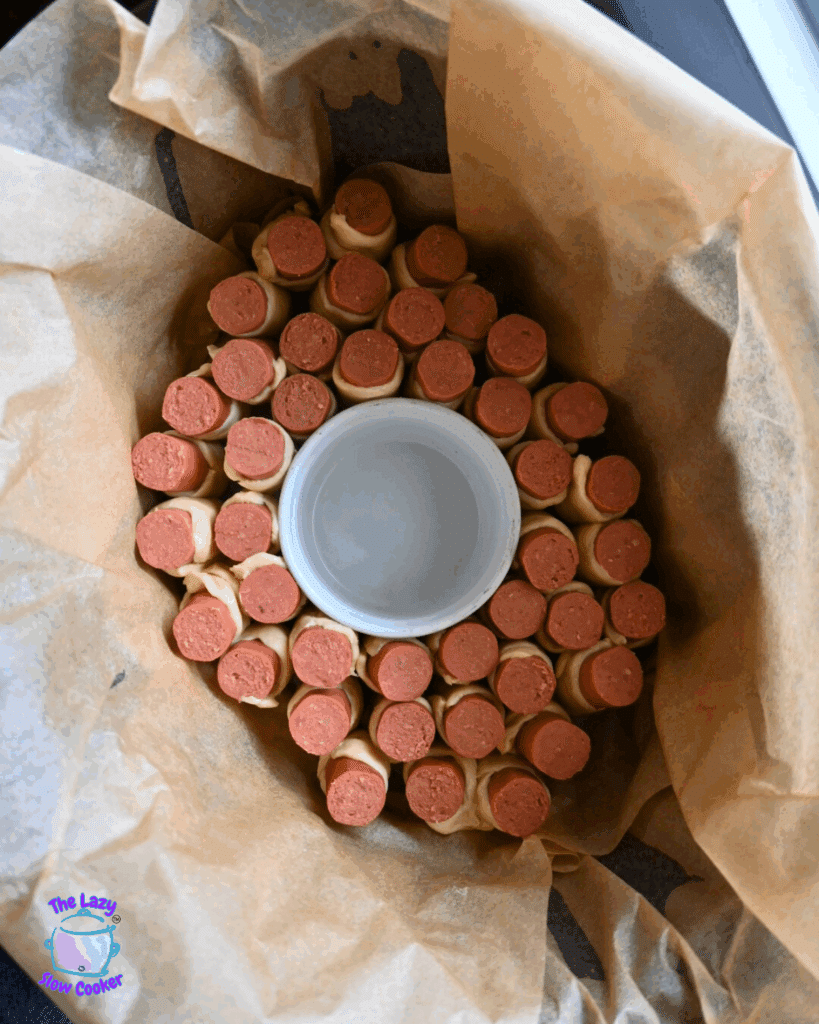 Uncooked hot dog pull apart in a a slow cooker with an empty white ramekin in the middle.