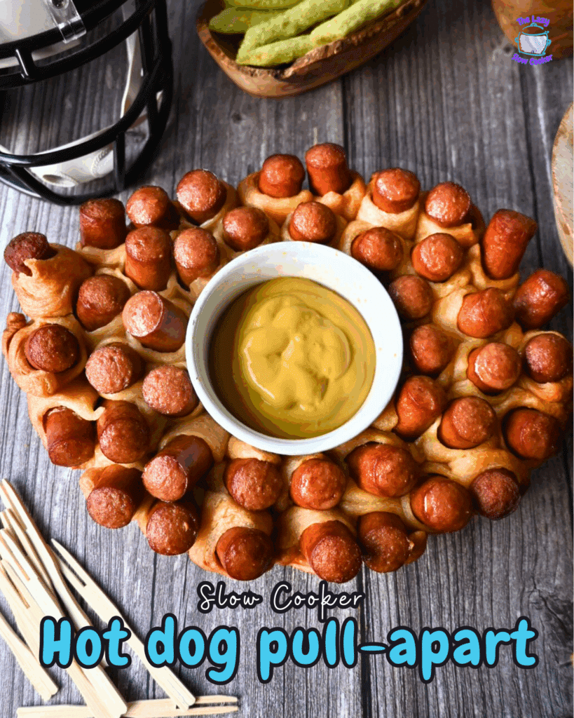 Oval slow cooker-shaped pull-apart bread with mini hot dogs baked in, arranged around a central bowl of yellow mustard dipping sauce, set on a wooden surface.