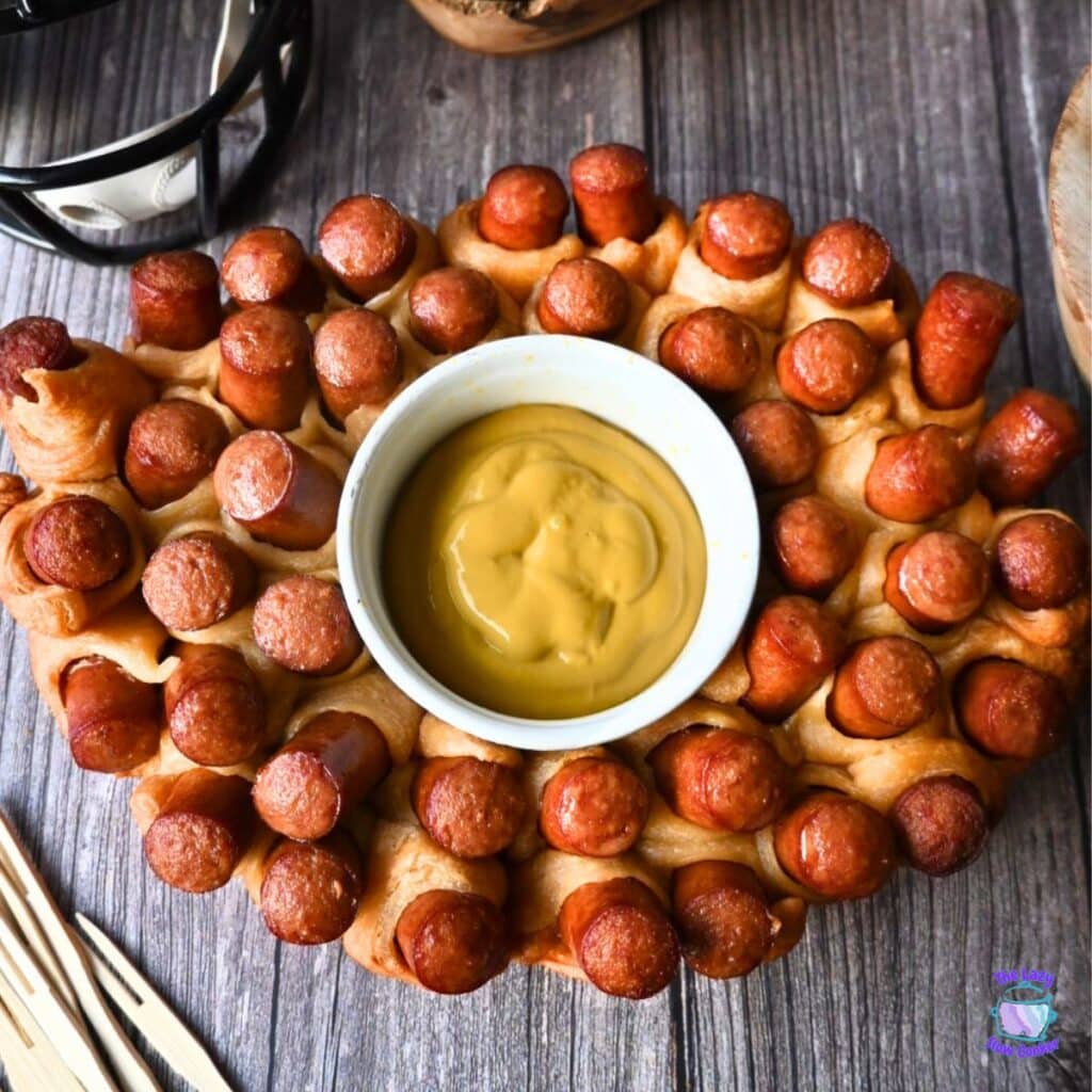 Oval slow cooker-shaped pull-apart bread with mini hot dogs baked in, arranged around a central bowl of yellow mustard dipping sauce