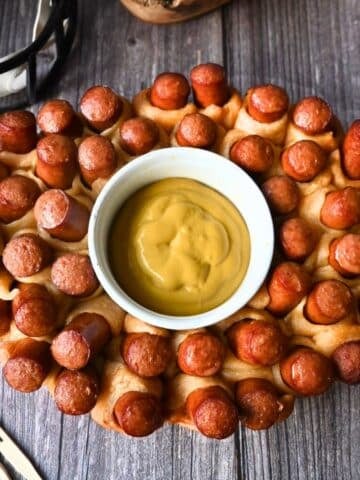 Oval slow cooker-shaped pull-apart bread with mini hot dogs baked in, arranged around a central bowl of yellow mustard dipping sauce