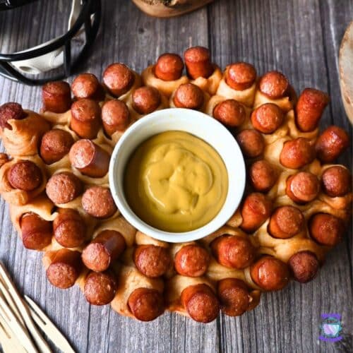 Oval slow cooker-shaped pull-apart bread with mini hot dogs baked in, arranged around a central bowl of yellow mustard dipping sauce