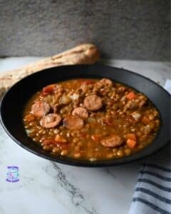 A black bowl filled with lentil soup and sliced sausage sits on a marble surface, with a piece of bread in the background and a striped cloth to the side.