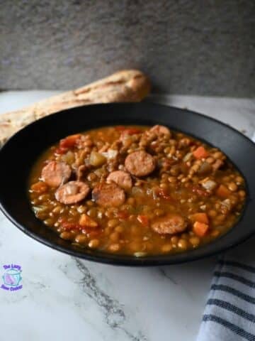 A black bowl filled with lentil soup and sliced sausage sits on a marble surface, with a piece of bread in the background and a striped cloth to the side.