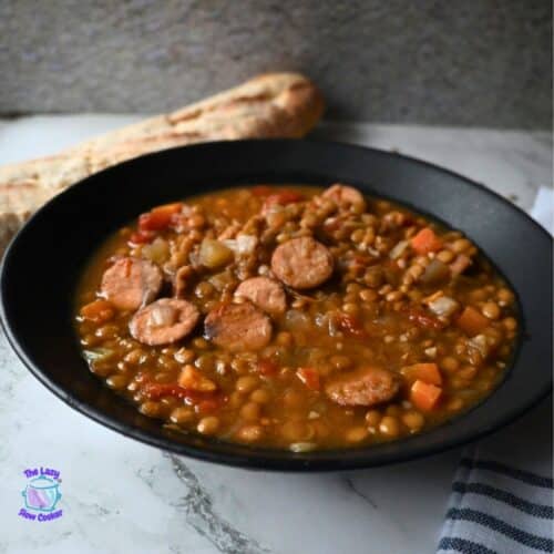 A black bowl filled with lentil soup and sliced sausage sits on a marble surface, with a piece of bread in the background and a striped cloth to the side.