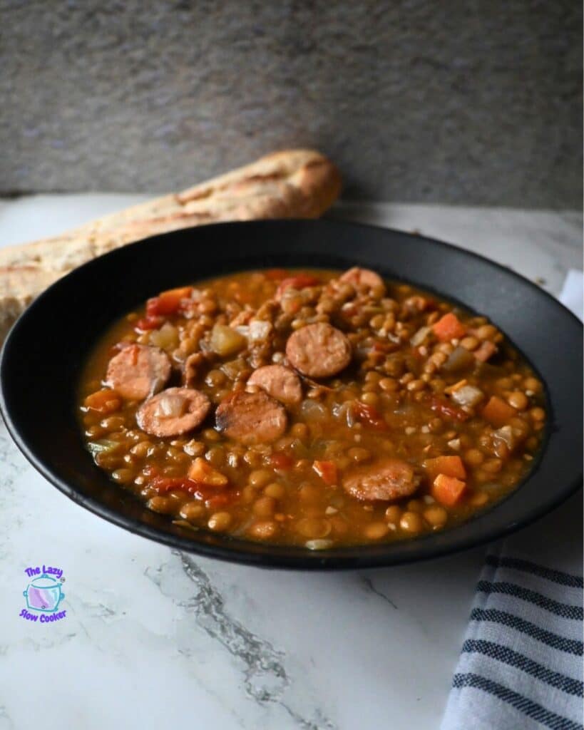 A black bowl filled with lentil soup and sliced sausage sits on a marble surface, with a piece of bread in the background and a striped cloth to the side.
