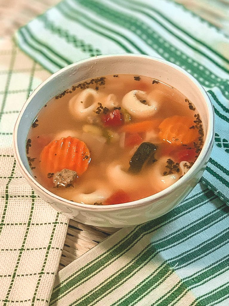 a white bowl filled with soup that has tortellini, veggies, and spices floating