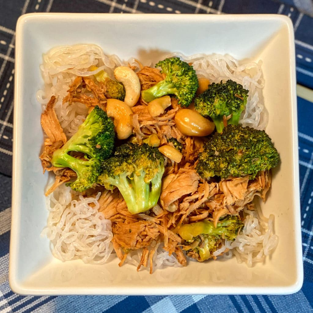 Looking down on a square white bowl filled with broccoli, chicken and cashew nuts over cellophane noodles
