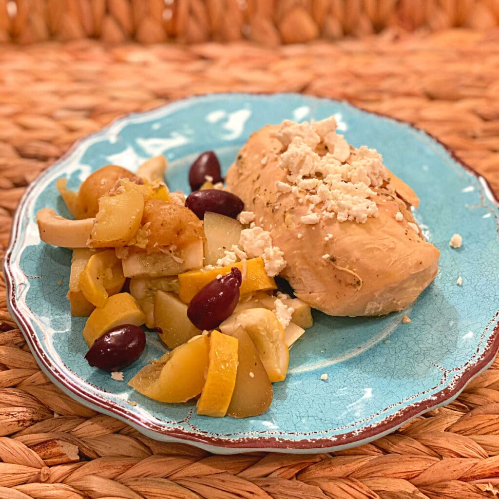 Turquoise plate with chicken breast topped with feta cheese, kalamata olives, and cooked veggies