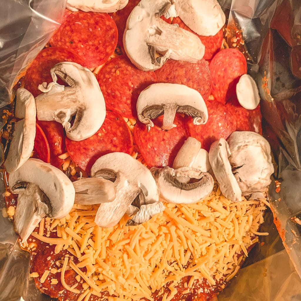 Top view of a crockpot filled with uncooked chicken, sauce, shredded cheese, pepperoni and mushrooms