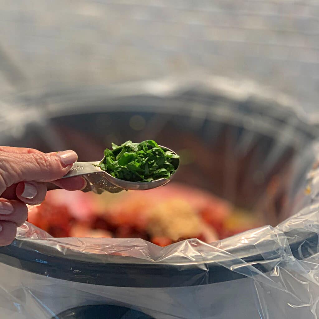 a measuring spoon full of freshly chopped basil being held over a blurry image of a crockpot in the background