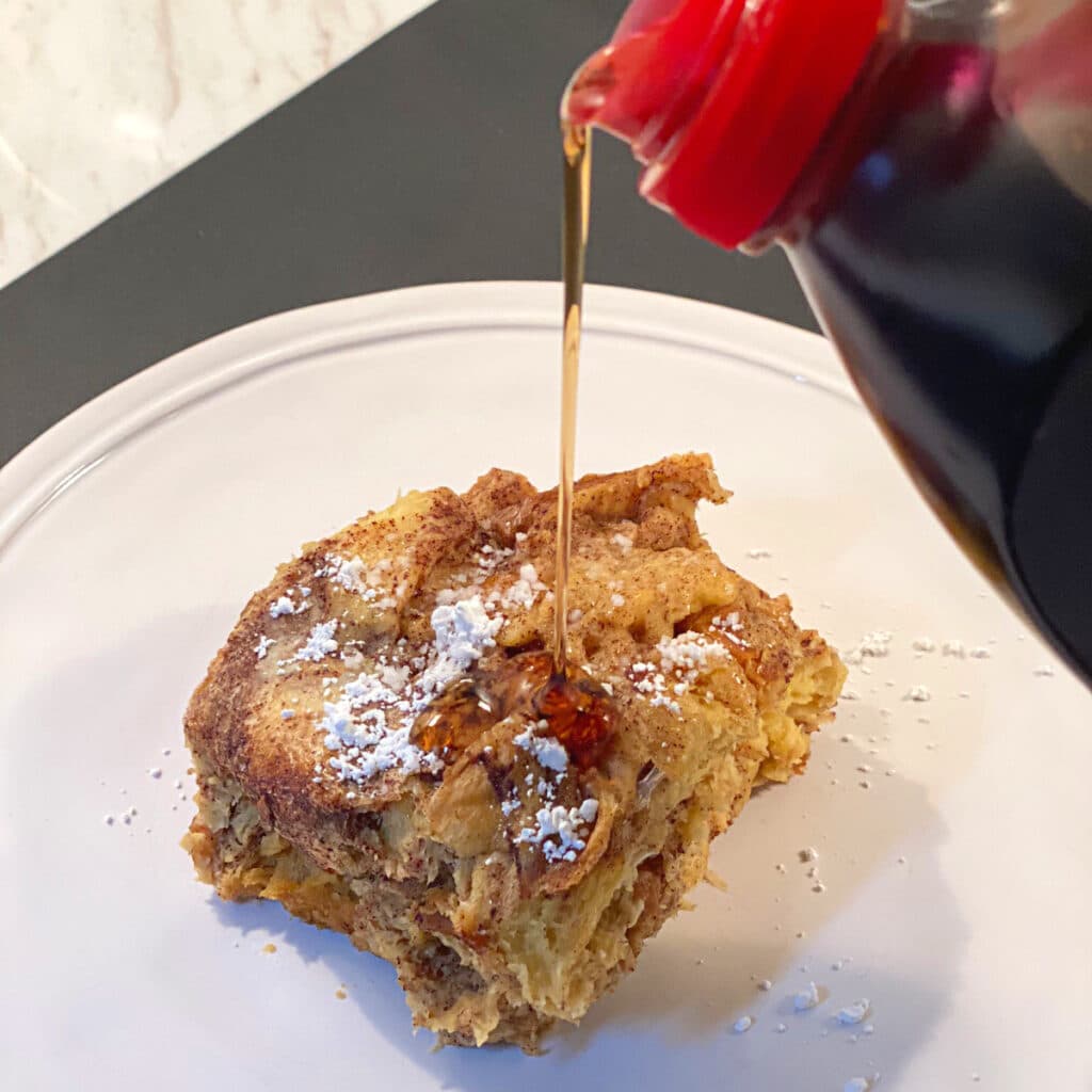 Syrup being poured over a piece of french toast on a round white plate