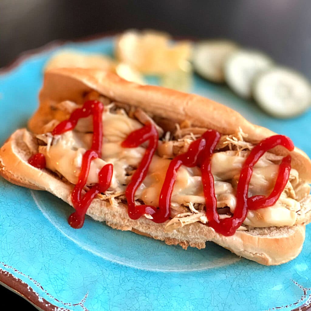 turquoise plate with finished sandwich drizzled with ketchup with pickles in the bakcground 