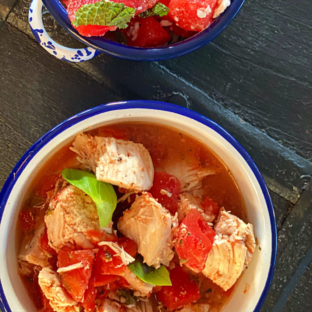 top view of chicken with tomato and basil in a round bowl with watermelon salad in background