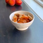a side view of peach cobbler in a round white bowl. blurred whole peaches inthe background