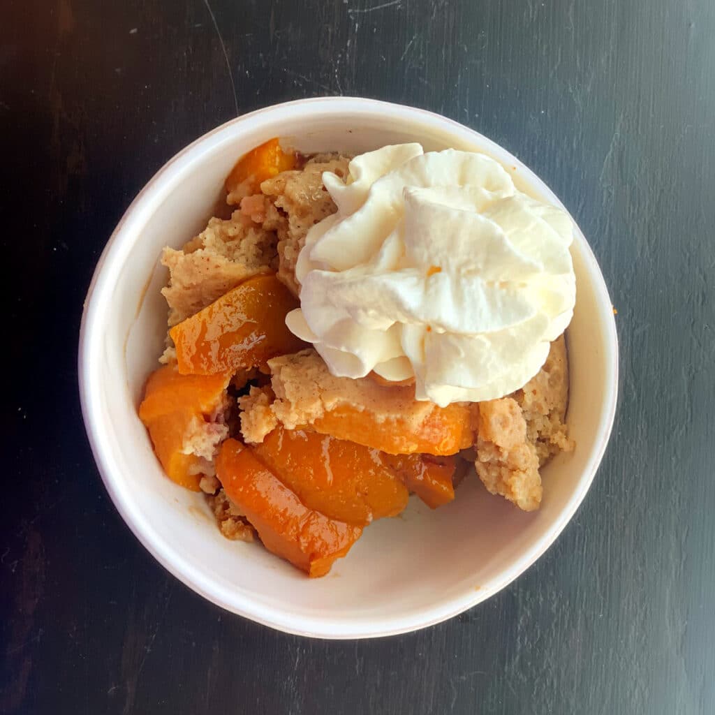 Looking down on finished peach dish topped with whipped cream in a round, white bowl on a black table