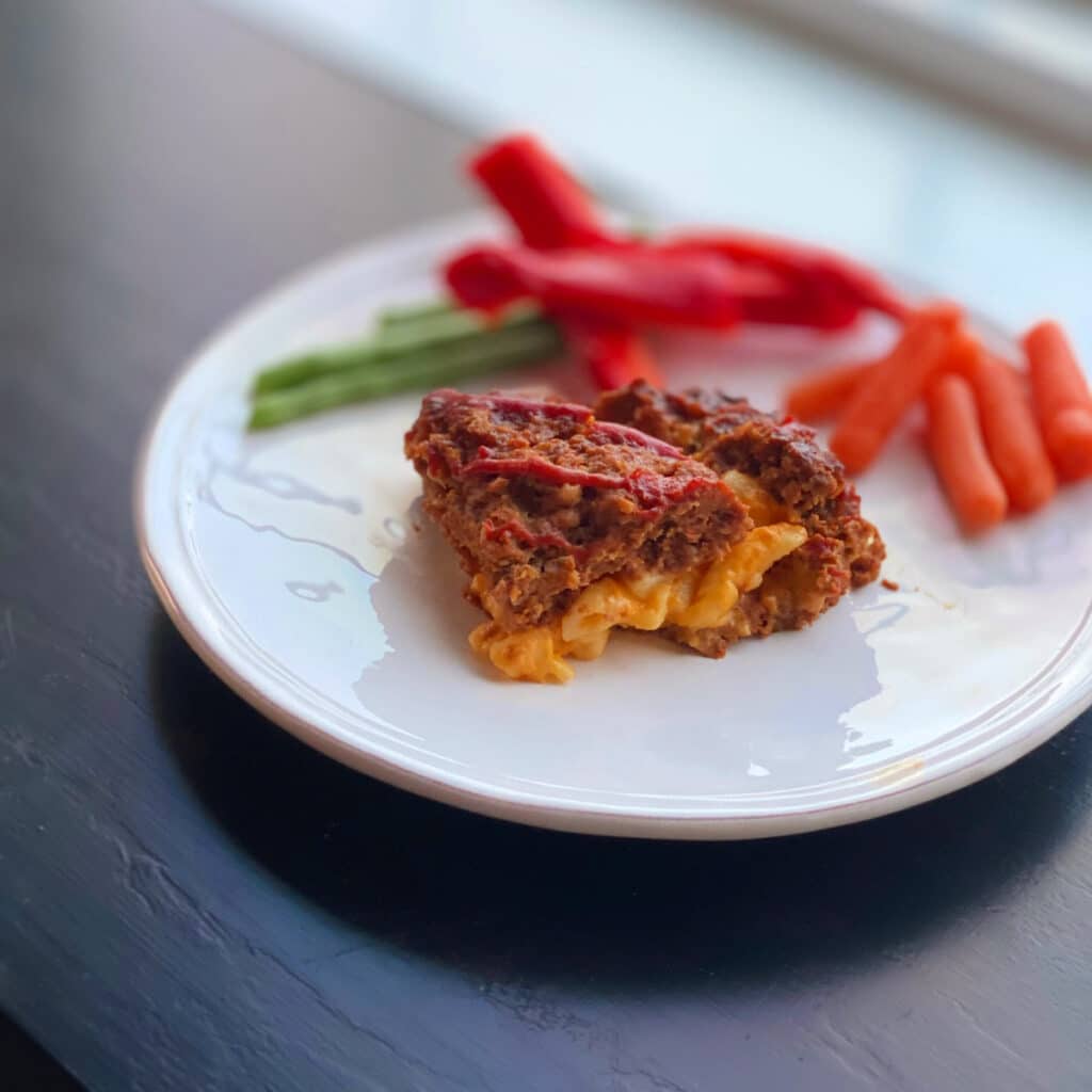 finished mac and cheese stuffed meatloaf with red peppers, carrots and green beans in the background on a round white plate