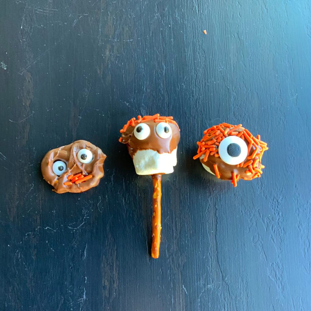 a small pretzel, a marshmallow and a marshmallow on a pretzel stick dipped in milk chocolate and decorated with orange jimmies and candy eyes