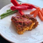 macaroni and cheese stuffed meatloaf on a round white place with colorful veggies in the background