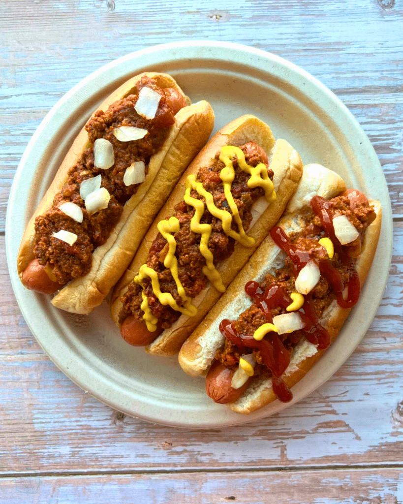 three hot dogs on a plate topped with chili and other toppings