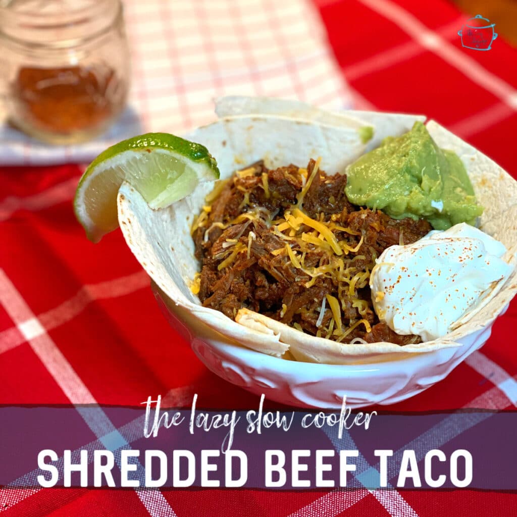 shredded beef taco in a tortilla bowl with guacamole and sour cream and topped with a little shredded cheese