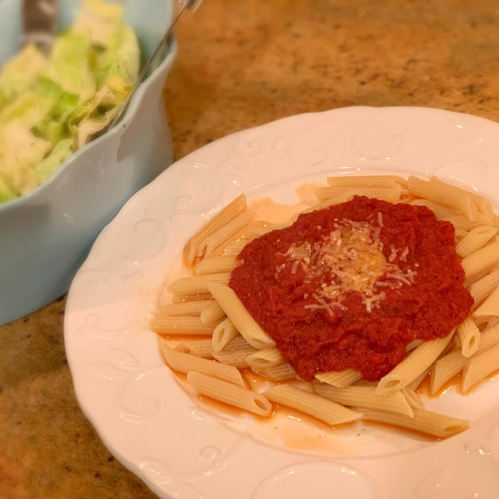 a round white plate filled with penne pasta, sauce and topped with some melted parmesan cheese. A blurred salad is in the background